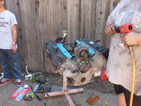 2003 Ford Crown Victoria - worker runs a pneumatic tool on the exposed V8 as a young onlooker stands by the fence