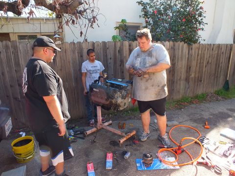 2003 Ford Crown Victoria - Three workers confer over the V8 on its stand, with part boxes and a yellow bucket on the concrete