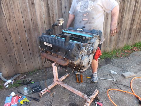 2003 Ford Crown Victoria - Side view of the bare V8 on its timber stand, exhaust manifold exposed, tools spread on the ground