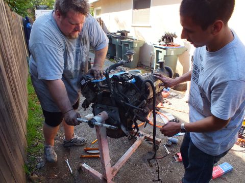 2003 Ford Crown Victoria - man and a younger helper handle the V8 on its stand near green trash bins during the rebuild