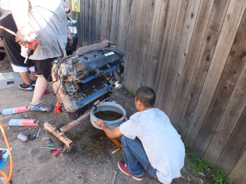 2003 Ford Crown Victoria - child steadies a bucket catching dark oil from the V8 on its stand as adults work on the left