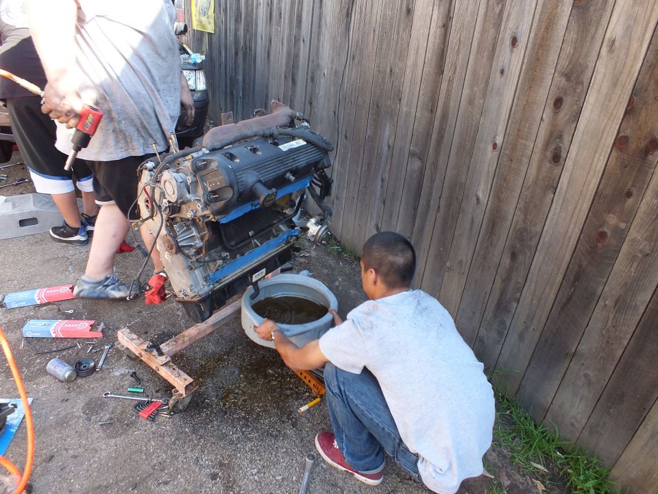 Young boy holding a bucket of dark fluid beside a V8 engine on a wooden stand against a wood fence