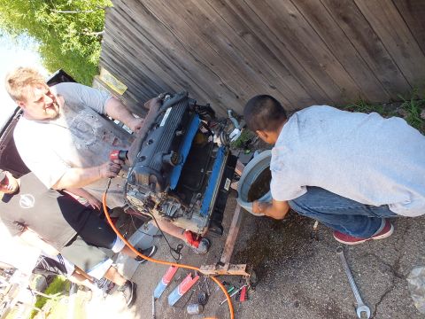 2003 Ford Crown Victoria - Overhead angle of the V8 on its timber stand with a man using an air tool and a boy crouched beneath