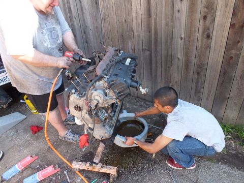 2003 Ford Crown Victoria - worker runs an air tool on the V8 while a helper crouches with a drain pan; CA plate partly visible left