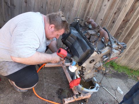 2003 Ford Crown Victoria - Worker drives an air tool into the bare V8 block on its timber stand, drain pan catching fluid below