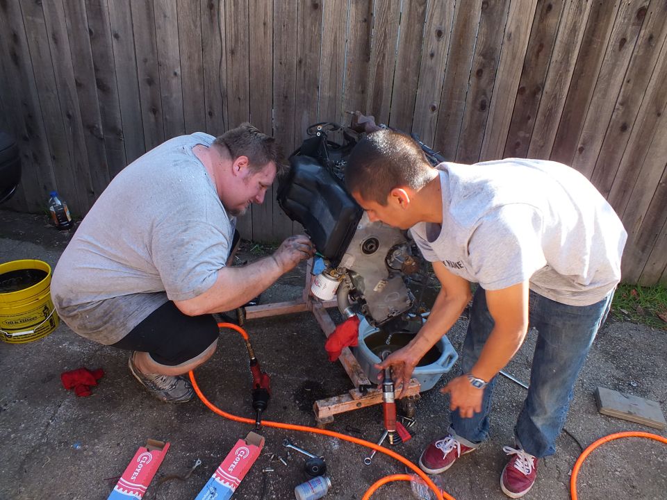 Two men crouched disassembling a removed engine on wood blocks against a backyard fence