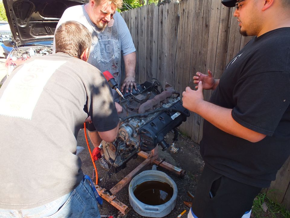 Three men working an impact wrench on a removed engine over a drain pan outdoors