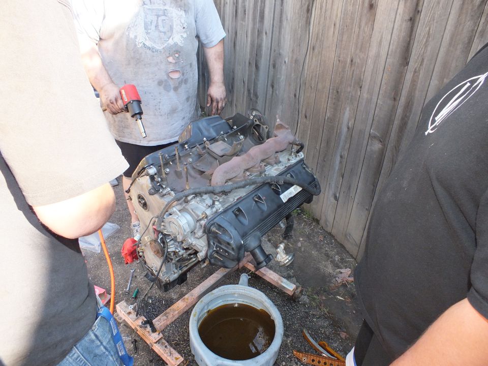 Two men using an impact wrench on a removed engine staged over an oil pan