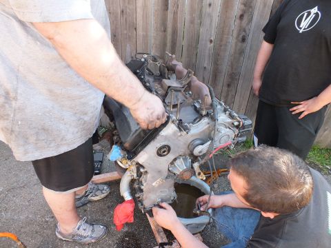 2003 Ford Crown Victoria - Mechanics working a removed engine over a drain pan, onlooker in a V-logo shirt at right