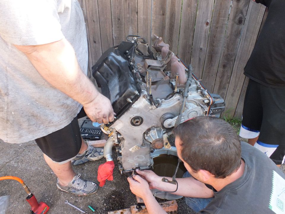 Two men removing components from a stripped engine block over an oil drain pan