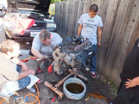2003 Ford Crown Victoria - Driveway engine teardown with helpers; SUV hood raised in background and tools spread on ground