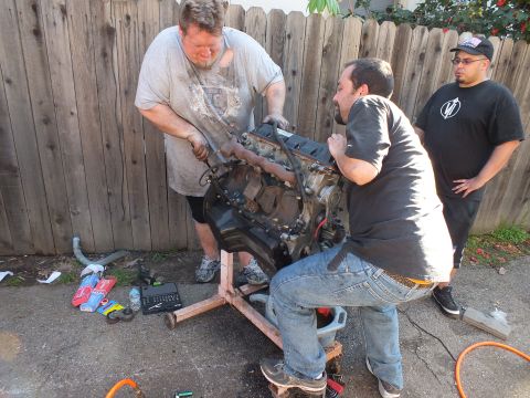 2003 Ford Crown Victoria - Men hoisting a bare engine on a wood frame, hand tools and sockets scattered on driveway