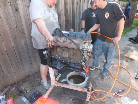 2003 Ford Crown Victoria - Engine perched on a wooden frame, one helper holding an orange air hose, oil pan underneath