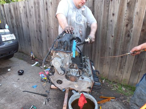 2003 Ford Crown Victoria - Greasy mechanic leans over an upended engine on a cradle, parts and rags on the pavement