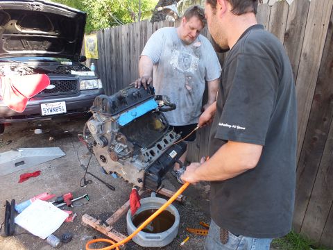 2003 Ford Crown Victoria - Pair working an engine on a cradle, SUV hood raised behind, shirt reads rebuilding 40 years