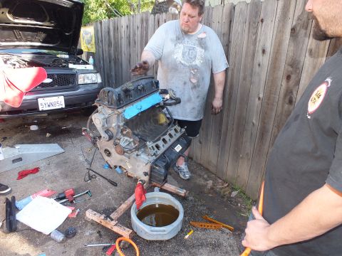 2003 Ford Crown Victoria - Engine on a wood frame in a driveway, SUV with raised hood and oil drain pan in the scene