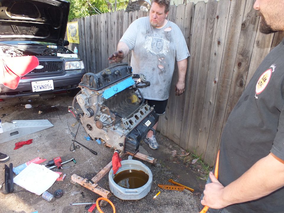 Engine on cradle between two men, dark SUV hood up, California plate 5YWN854 visible