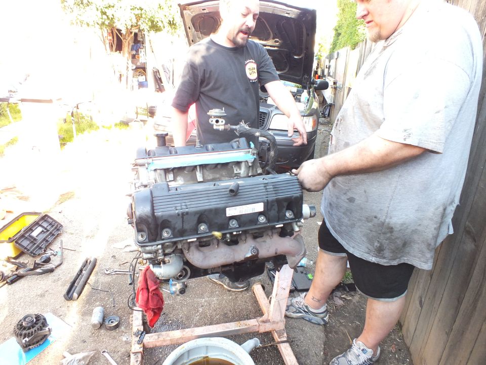 Two men beside engine with valve covers on cradle, vehicle with open hood behind
