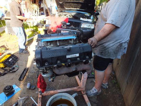 2003 Ford Crown Victoria - Bare engine on a wooden cradle near a raised SUV hood, hand tools strewn across pavement