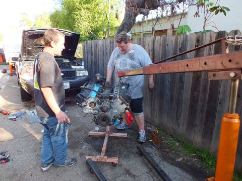 2003 Ford Crown Victoria - Engine on a wood frame beside a red shop crane, vehicle with raised hood in background