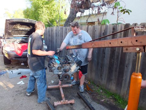 2003 Ford Crown Victoria - Pair maneuvering an engine on a wood frame next to a red hoist, dark car hood up behind