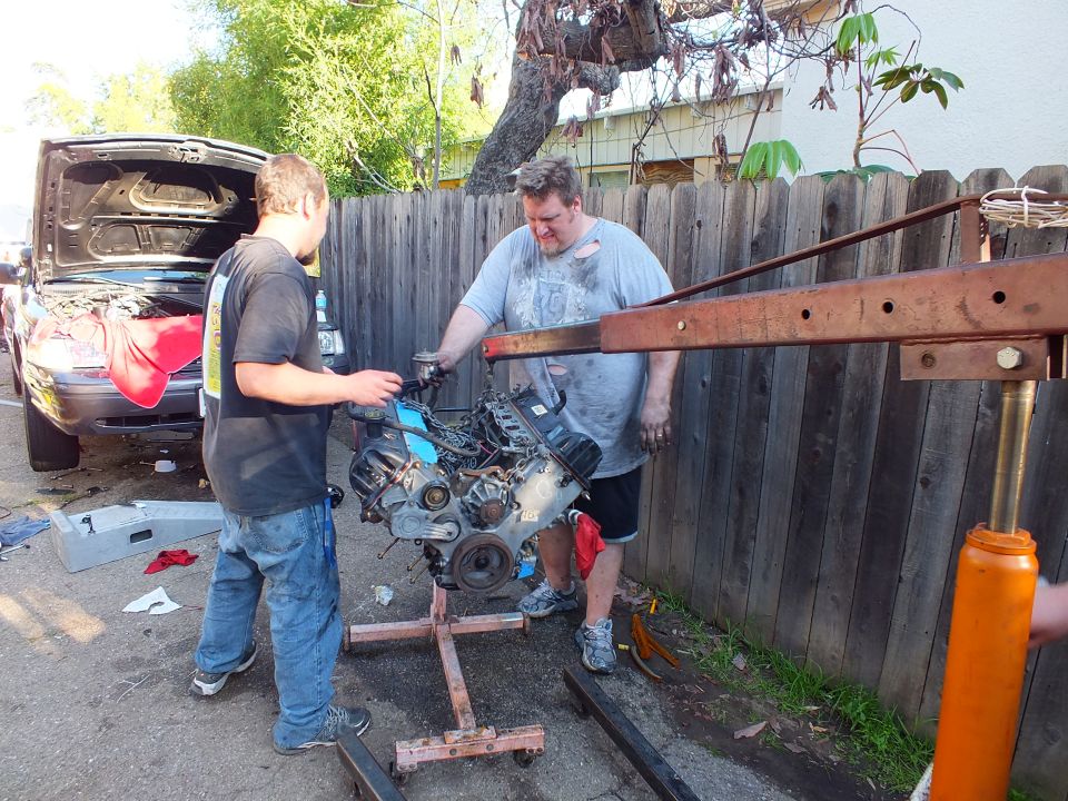 Two men lifting engine onto cradle beside red engine crane and dark car with open hood