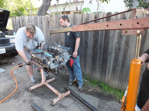 2003 Ford Crown Victoria - Mechanic turning a wrench on an engine beneath a red crane boom, helper standing alongside