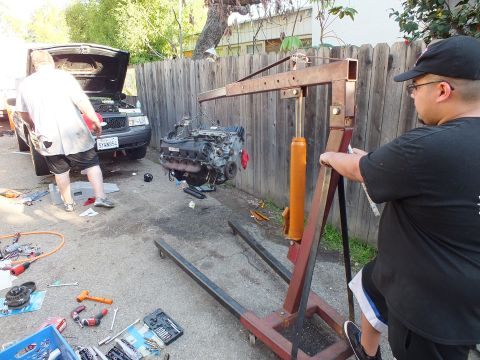 2003 Ford Crown Victoria - Engine slung from a red shop crane in a driveway as a man works the hoist, hood-up car left