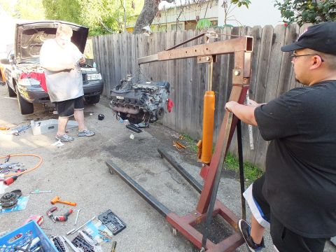 2003 Ford Crown Victoria - Bare engine lifted by a red folding shop crane near a fence, tool sets spread on the ground