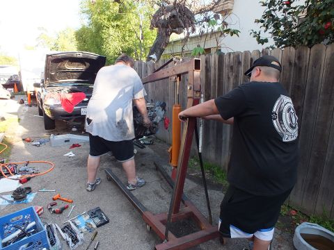 2003 Ford Crown Victoria - Engine staged by a red shop crane as two men work, hood-up vehicle and toolboxes in driveway