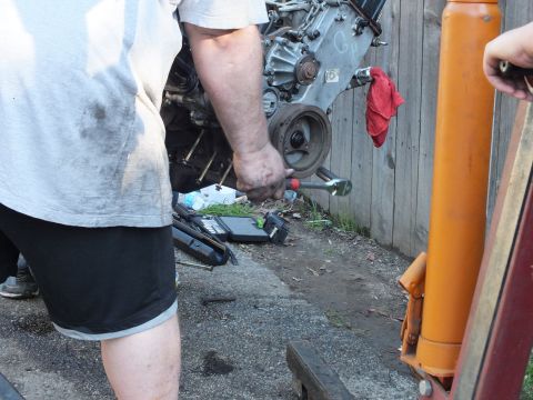 2003 Ford Crown Victoria - Hands working a ratchet on a bare engine against a fence, orange hoist cylinder at right