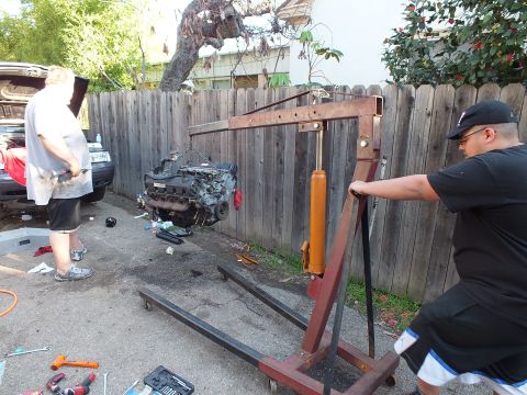 2003 Ford Crown Victoria - Man hauling the lift strap of a red shop crane to raise an engine, hood-up car at far left