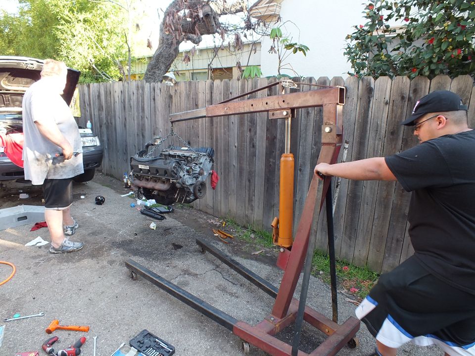 Man pulling chain on red engine crane lifting engine in driveway near wooden fence