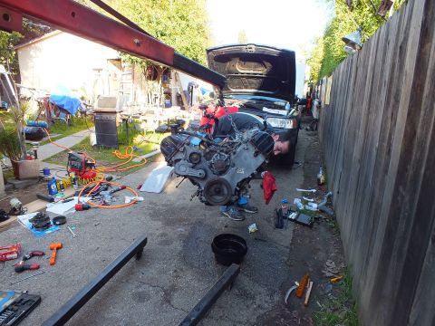2003 Ford Crown Victoria - V6 engine hangs from a hoist arm beside a dark SUV with hood raised; tools and air hoses cover the driveway.