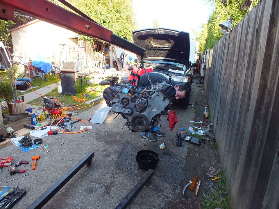 Engine on hoist in front of dark SUV with open hood in cluttered driveway