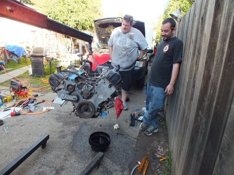 2003 Ford Crown Victoria - Two men stand by a removed engine hanging from a hoist; wrenches, hoses and a drain pan litter the driveway.