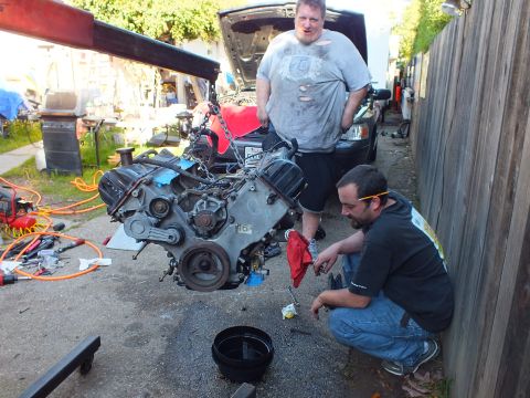 2003 Ford Crown Victoria - kneeling worker tends to a chain-hoisted engine as a second man looks on in a fenced driveway.