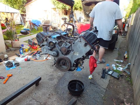 2003 Ford Crown Victoria - Removed engine block, marked V6 in chalk, hangs from a hoist amid scattered parts and tools.