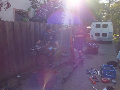2003 Ford Crown Victoria - Two men maneuver an engine hoist near a wood fence under heavy sun flare; a white van sits in the driveway.