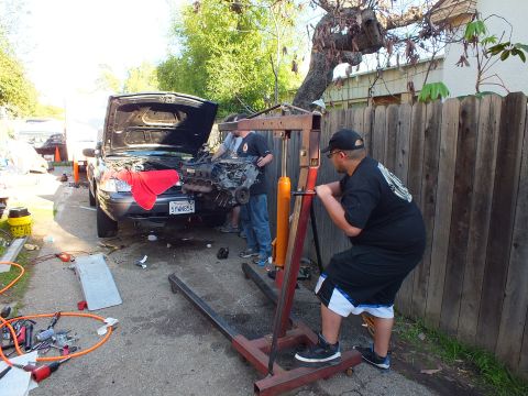 2003 Ford Crown Victoria - worker leans into an engine crane wheeled toward a dark SUV, hood up and a red cloth draped over the fender.