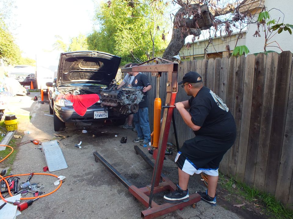 Man pushes engine hoist toward dark SUV with open hood and red cloth on fender