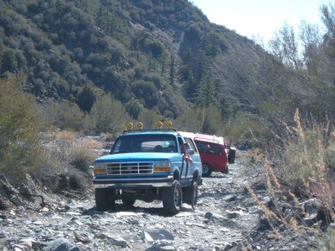 1988 Ford Bronco - A blue SUV with auxiliary lights on its roof rack navigates a rocky, off-road trail, followed by a red SUV.
