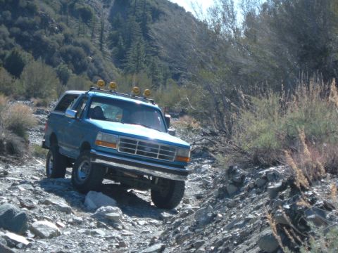 1988 Ford Bronco - Blue Ford Bronco navigates a rough, rocky terrain on a sunny day.