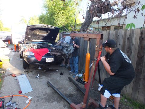 2003 Ford Crown Victoria - engine crane is set up at the nose of a hood-up SUV, California plate 5YWN854 visible, as workers gather.
