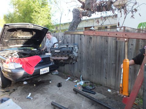 2003 Ford Crown Victoria - man grins behind an engine suspended over a dark SUV's bay; California plate 5YWN854 and red fender cloth show.