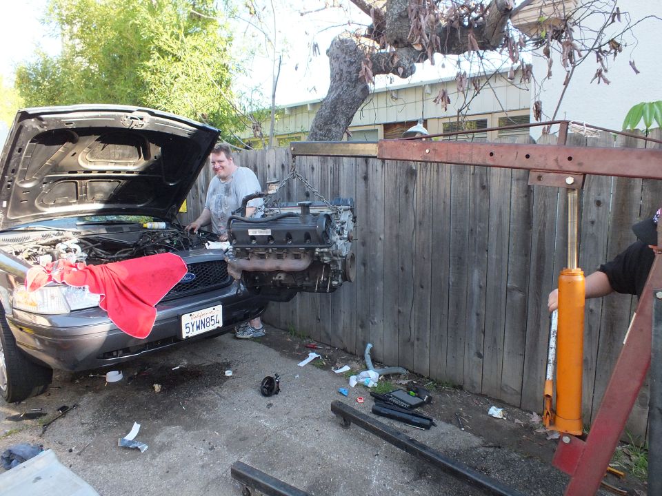 Man smiles beside engine lifted on chain over front of dark SUV with red cloth