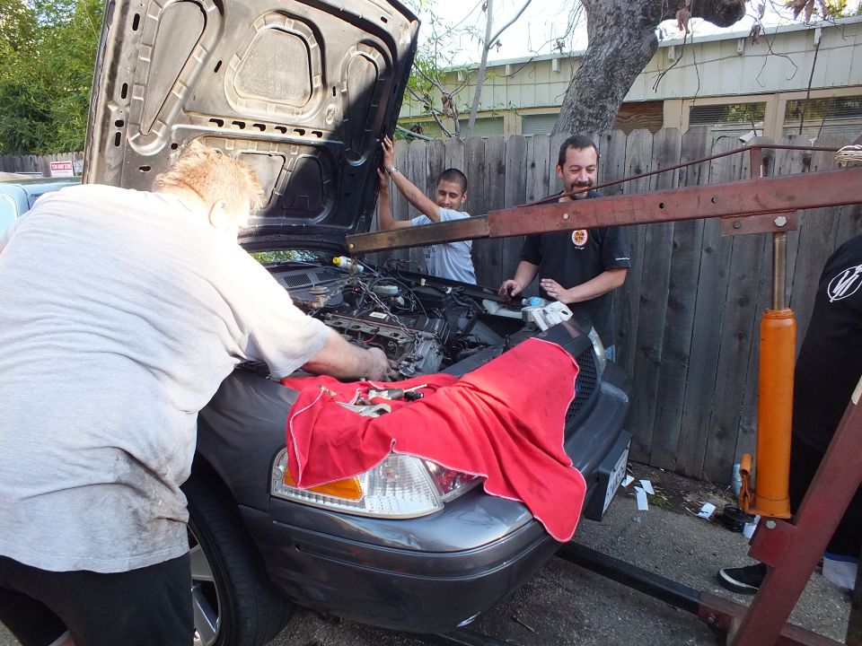 Men lower engine into front of gray SUV with red cloth over fender