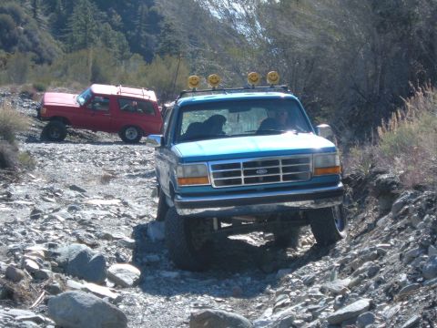 1988 Ford Bronco - Two Ford Broncos tackle a challenging, rocky off-road path under a clear sky.