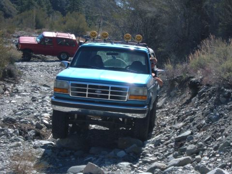 1988 Ford Bronco - A blue Ford Bronco navigates a , rocky trail, with another red SUV visible in the background.