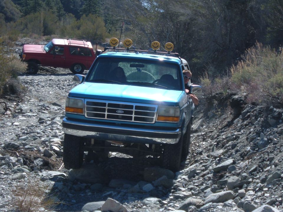 Blue Ford Bronco with smiley face lights on roof rack, driving on rocky terrain
